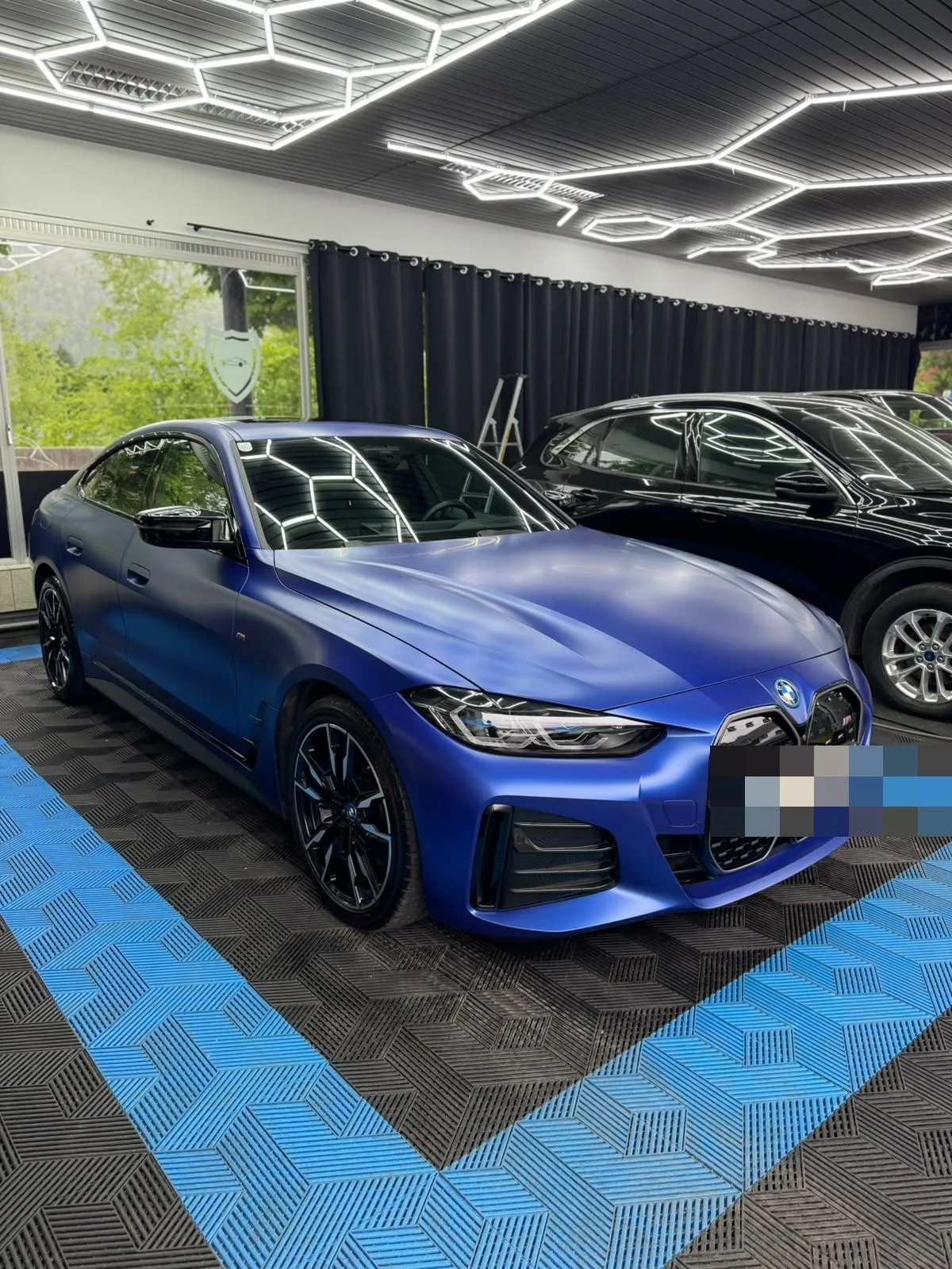 BMW i4 in Mattblau in der Halle von Saff Car Lounge in Kufstein nach der Aufbereitung, Hexagon-Leuchten spiegeln sich im Lack